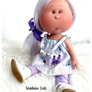 May include: A white doll with long, white hair wearing a white dress with a purple and blue pattern and purple leggings. The doll is wearing white shoes with brown soles. The doll is sitting on a white fluffy surface. The text "TickleBebe Dolls" is visible in the bottom center of the image.
