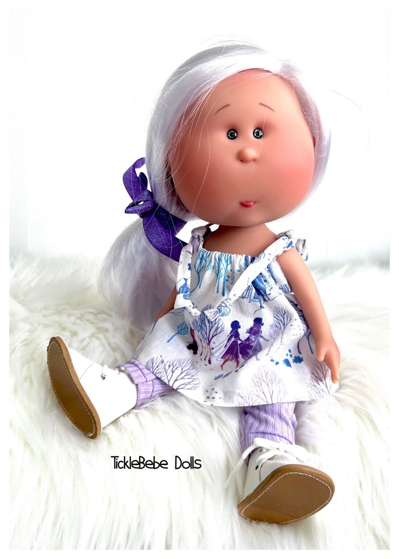 May include: A white doll with long, white hair wearing a white dress with a purple and blue pattern and purple leggings. The doll is wearing white shoes with brown soles. The doll is sitting on a white fluffy surface. The text "TickleBebe Dolls" is visible in the bottom center of the image.