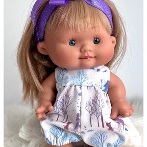 May include: A small doll with blonde hair wearing a purple headband with a bow and a white dress with a blue and purple winter scene print. The doll is sitting on a white fluffy surface.