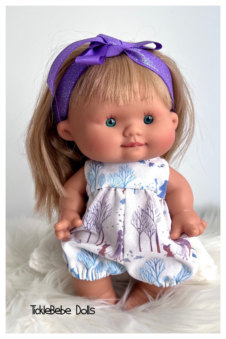 May include: A small doll with blonde hair wearing a purple headband with a bow and a white dress with a blue and purple winter scene print. The doll is sitting on a white fluffy surface.