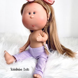 May include: A doll with long blonde hair wearing a pink headband with white polka dots and a purple ribbed outfit. The doll is sitting on a white fluffy surface. The text "TickleBabe Dolls" is visible in the bottom left corner of the image.