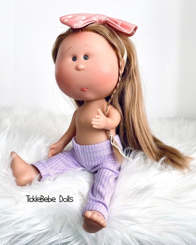 May include: A doll with long blonde hair wearing a pink headband with white polka dots and a purple ribbed outfit. The doll is sitting on a white fluffy surface. The text "TickleBabe Dolls" is visible in the bottom left corner of the image.
