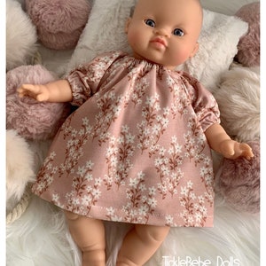 May include: A baby doll wearing a pink floral dress, lying on a white fluffy surface. The dress features a white floral pattern with brown stems. The doll is surrounded by pink and white fluffy pom-poms and a white pillow. The text "TickleBebe Dolls" is visible.