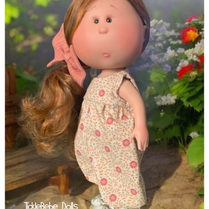 May include: A handmade doll with long brown hair, wearing a floral print dress and silver shoes with yellow soles. The doll is standing on a rock with a wooden bench behind it. The doll is looking to the right of the image. The text "TickleBebe Dolls" is visible in the bottom left corner of the image.