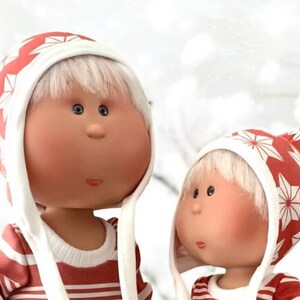 May include: Two dolls with rosy cheeks and blue eyes. They wear red and white striped shirts and matching hats with a red and white star pattern. The dolls have white hair and are set against a blurred, snowy background.