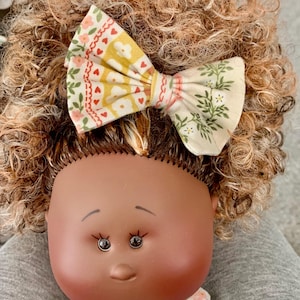 May include: A doll with brown skin and curly brown hair wearing a floral print bow. The doll has large brown eyes and a sweet expression.