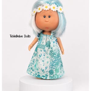 May include: A doll with light blue hair, a white daisy flower crown, and freckles on its face. The doll wears a sleeveless dress featuring a teal and white floral pattern. The text TickleBebe Dolls is printed in black on the left side of the doll.