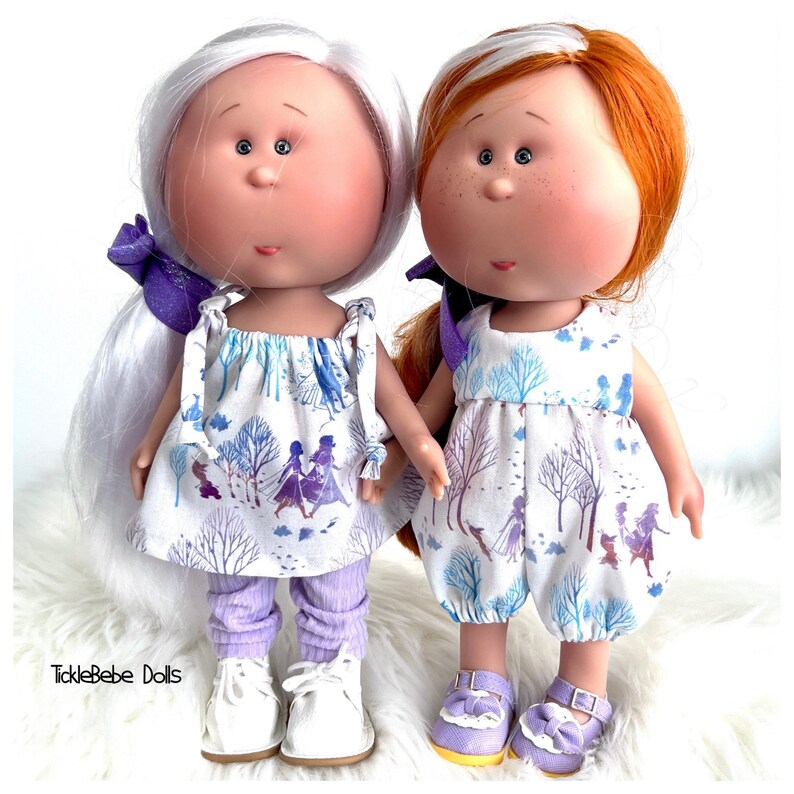 May include: Two handmade dolls with white dresses featuring a winter scene and purple accents. The dolls have white and red hair, and one is wearing purple pants and white shoes, while the other is wearing purple shoes.