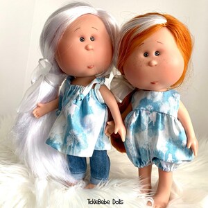 May include: Two handmade dolls with painted faces and long hair. One doll has white hair and is wearing a blue and white dress with denim pants. The other doll has red hair and is wearing a blue and white dress. The dolls are standing on a white fluffy surface. The text "TickleBabe Dolls" is visible at the bottom of the image.