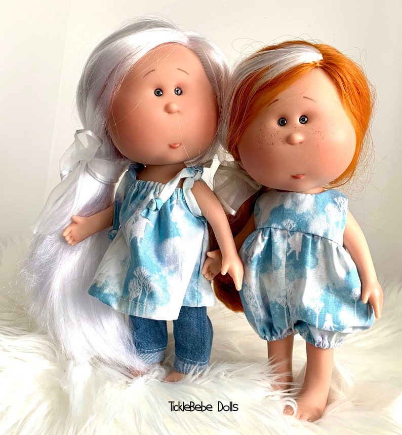 May include: Two handmade dolls with painted faces and long hair. One doll has white hair and is wearing a blue and white dress with denim pants. The other doll has red hair and is wearing a blue and white dress. The dolls are standing on a white fluffy surface. The text "TickleBabe Dolls" is visible at the bottom of the image.