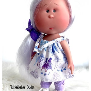 May include: A handmade doll with white hair, a purple bow, and a white dress with a blue and purple print of two girls walking in a snowy forest. The doll is wearing purple leggings and white shoes. The doll is standing on a white fluffy surface. The text "TickleBebe Dolls" is visible in the bottom left corner of the image.