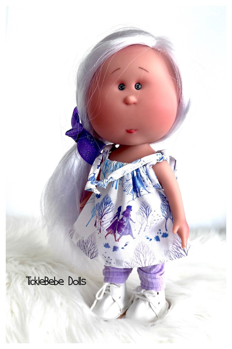 May include: A handmade doll with white hair, a purple bow, and a white dress with a blue and purple print of two girls walking in a snowy forest. The doll is wearing purple leggings and white shoes. The doll is standing on a white fluffy surface. The text "TickleBebe Dolls" is visible in the bottom left corner of the image.