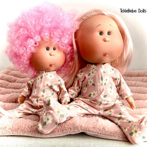 May include: Two dolls wearing pink and white floral pajamas. The doll on the left has pink curly hair and the doll on the right has long blonde hair. The dolls are sitting on a pink fuzzy blanket. Ticklebebe Dolls is written in the top right corner of the image.