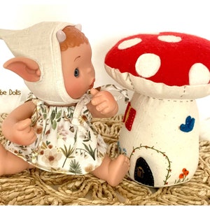 May include: A small elf doll with pointed ears wearing a white hat and a floral dress sits next to a red and white mushroom house with a blue bird and a small door.