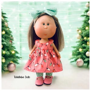 May include: A handmade doll wearing a pink dress with a Christmas lights pattern, a green headband with white polka dots, and pink shoes. The doll is standing in front of a green background with Christmas trees.