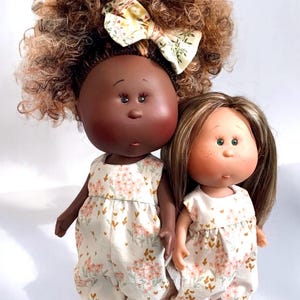 May include: Two handmade dolls wearing floral print overalls. The doll on the left has brown skin and curly brown hair with a yellow and green bow. The doll on the right has light skin and straight brown hair.