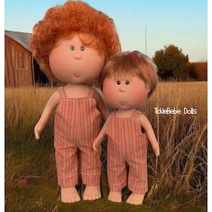 May include: Two red-haired dolls wearing matching red and white striped overalls. The dolls are standing in a field of tall grass.
