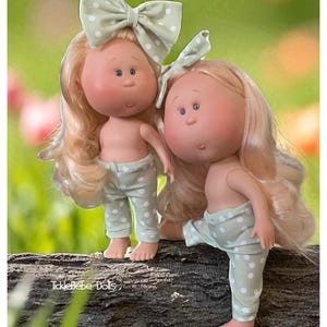 May include: Two handmade dolls with blonde hair, rosy cheeks, and blue eyes. They wear light green polka dot pants and matching bows. The dolls stand on a log against a blurred green background. The text "TickleBebe Dolls" is visible.