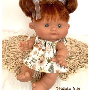 May include: A doll with red hair styled in two buns, blue eyes, and a floral print dress. The dress features a pattern of flowers, mushrooms, and leaves. The doll is sitting on a woven mat. The text "TickleBebe Dolls" is visible.