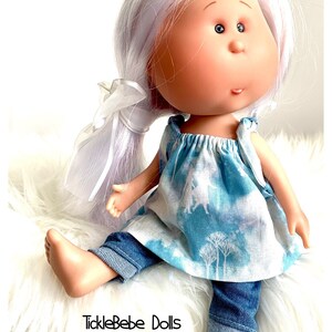 May include: A doll with long, white hair tied back with a white ribbon. The doll is wearing a blue and white patterned dress and blue denim jeans. The doll is sitting on a white fluffy surface. The text "TickleBebe Dolls" is visible in the lower left corner of the image.