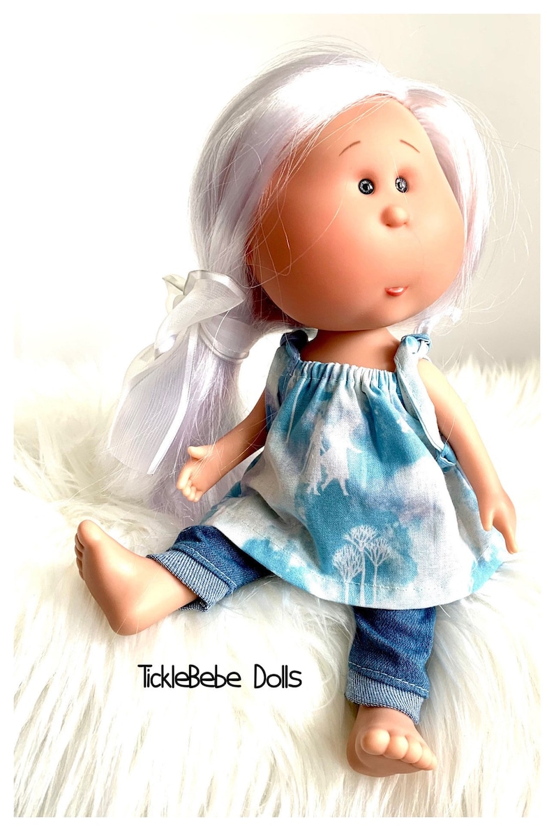 May include: A doll with long, white hair tied back with a white ribbon. The doll is wearing a blue and white patterned dress and blue denim jeans. The doll is sitting on a white fluffy surface. The text "TickleBebe Dolls" is visible in the lower left corner of the image.
