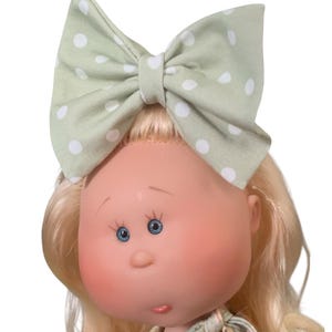 May include: A doll with blonde hair and blue eyes wearing a large sage green bow with white polka dots. The doll has rosy cheeks and a floral patterned dress. The text "TickleBebe Dolls" is visible at the bottom.