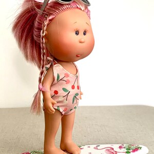 May include: A pink haired doll wearing sunglasses and a pink and green cherry print swimsuit stands on a white surfboard with a pink flamingo design.