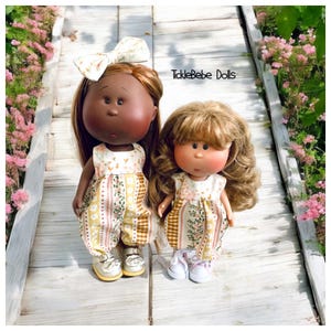 May include: Two handmade dolls wearing floral print dresses. The doll on the left has brown skin and a yellow and white striped dress. The doll on the right has blonde hair and a brown and white dress. The dolls are standing on a wooden walkway with flowers in the background. Ticklebebe Dolls is written on the walkway.