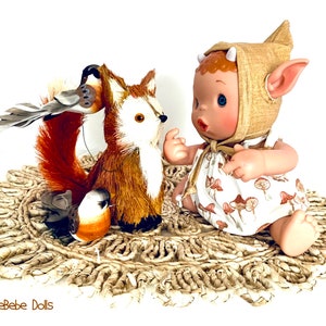 May include: A small, brown and white doll with pointed ears and a tan crocheted hat sits on a woven mat. The doll is looking at a brown and white fox figurine with a bird perched on its head. The fox is sitting on the mat with a small bird figurine beside it.