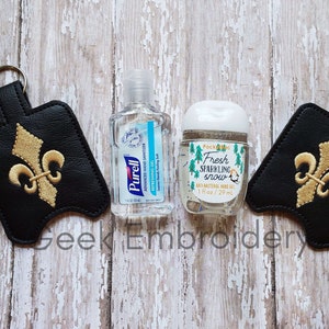 May include: Two black leather hand sanitizer holders with gold fleur-de-lis embroidery. Each holder has a key ring and is attached to a small bottle of hand sanitizer. One bottle is labeled "Purell" and the other is labeled "Fresh Sparkling Snow".