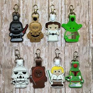 May include: Set of ten embroidered keychains featuring characters from Star Wars. The keychains are made of felt and have a variety of colors, including green, brown, white, black, and gold. The characters include Yoda, Chewbacca, Darth Vader, Princess Leia, and Boba Fett.