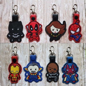 May include: Eight colorful keychains featuring different superhero characters. The characters include Deadpool, Black Panther, Spider-Man, Black Widow, Iron Man, Doctor Strange, Vision, and Scarlet Witch.