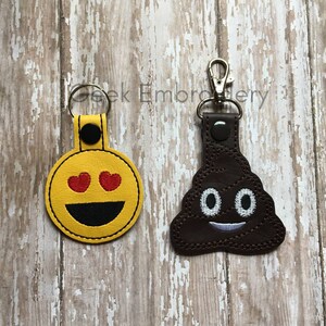 May include: Two embroidered keychains with different emoji faces. The first keychain is yellow with a smiley face with hearts for eyes. The second keychain is brown with a poop emoji with a smiley face.