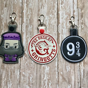 May include: Three Harry Potter themed keychains. The first keychain features a cartoon image of a wizard with a purple hat and glasses. The second keychain is a red and white circle with the text "The Bank of Gringotts". The third keychain is a black circle with the text "9 3/4".