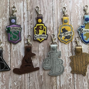 May include: Eight Harry Potter themed keychains with different house crests and characters. The keychains are made of leather and feature embroidered designs. The houses represented are Gryffindor, Hufflepuff, Slytherin, and Ravenclaw. The characters represented are the Sorting Hat, the Deathly Hallows symbol, and the initials "FB" and "HP".