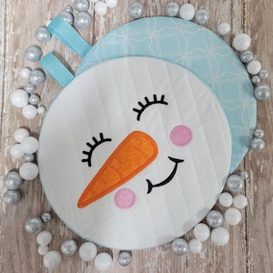 May include: A white snowman ornament with a blue and white patterned background. The snowman has a smiling face with black eyes, a carrot nose, and a pink blush.