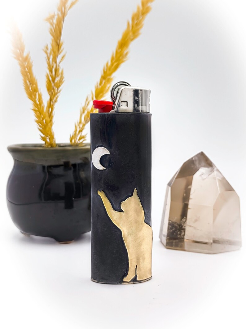 May include: Black lighter with a gold cat reaching for a silver crescent moon. The lighter has a red button and silver metal top. A small black pot with dried wheat and a clear crystal are in the background.
