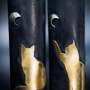 May include: Two cylindrical vases with a dark, textured finish. Each vase features a gold-colored cat silhouette and a silver crescent moon. One cat is seated, the other reaching up. A unique decorative item.