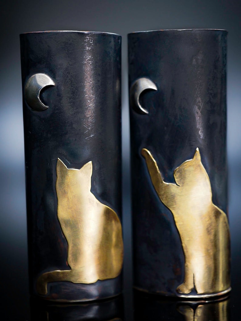May include: Two cylindrical vases with a dark, textured finish. Each vase features a gold-colored cat silhouette and a silver crescent moon. One cat is seated, the other reaching up. A unique decorative item.
