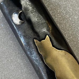 May include: A dark metal object with a gold cat silhouette and a silver crescent moon. The cat is sitting, and the moon is in the upper left corner. The metal has a textured appearance.