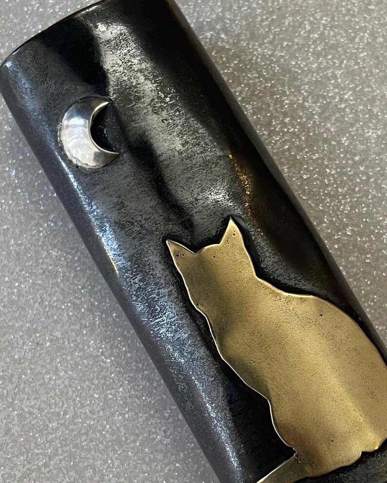 May include: A dark metal object with a gold cat silhouette and a silver crescent moon. The cat is sitting, and the moon is in the upper left corner. The metal has a textured appearance.