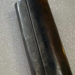 May include: Close-up of a cylindrical metal object with a dark, aged appearance. The object is divided into two sections by a vertical seam. The metal has a slightly textured surface and shows signs of wear, suggesting age or use.