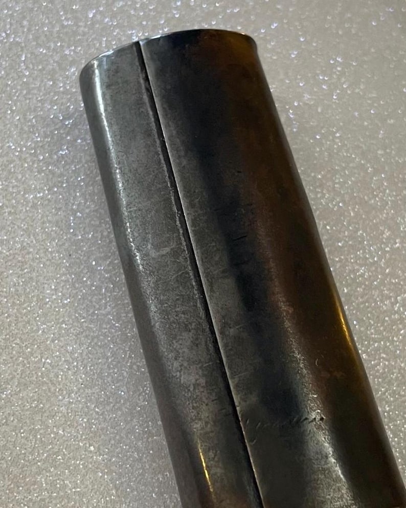 May include: Close-up of a cylindrical metal object with a dark, aged appearance. The object is divided into two sections by a vertical seam. The metal has a slightly textured surface and shows signs of wear, suggesting age or use.