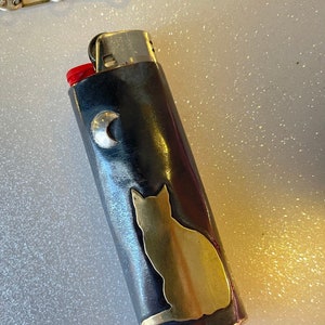 May include: A black and silver lighter with a gold cat silhouette and a silver crescent moon. The lighter has a red ignition button and a silver chain is visible in the background.