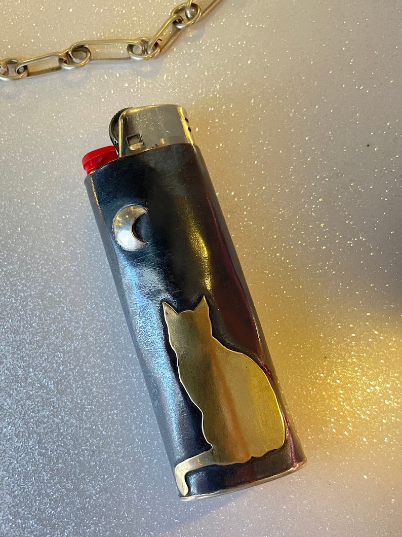 May include: A black and silver lighter with a gold cat silhouette and a silver crescent moon. The lighter has a red ignition button and a silver chain is visible in the background.