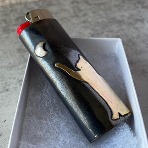 May include: A black lighter with a silver cat design and a silver crescent moon. The lighter has a red ignition button and is in a white box with white padding.