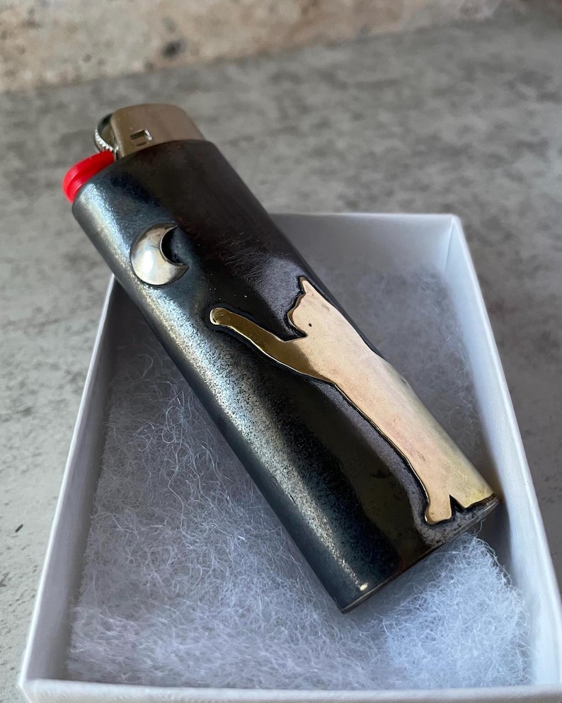 May include: A black lighter with a silver cat design and a silver crescent moon. The lighter has a red ignition button and is in a white box with white padding.