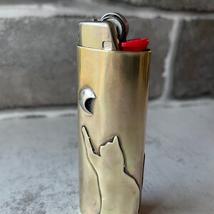 May include: A brass-colored lighter with a cat silhouette reaching for a crescent moon. The lighter has a red ignition button and a silver metal top. The design is etched into the brass casing.