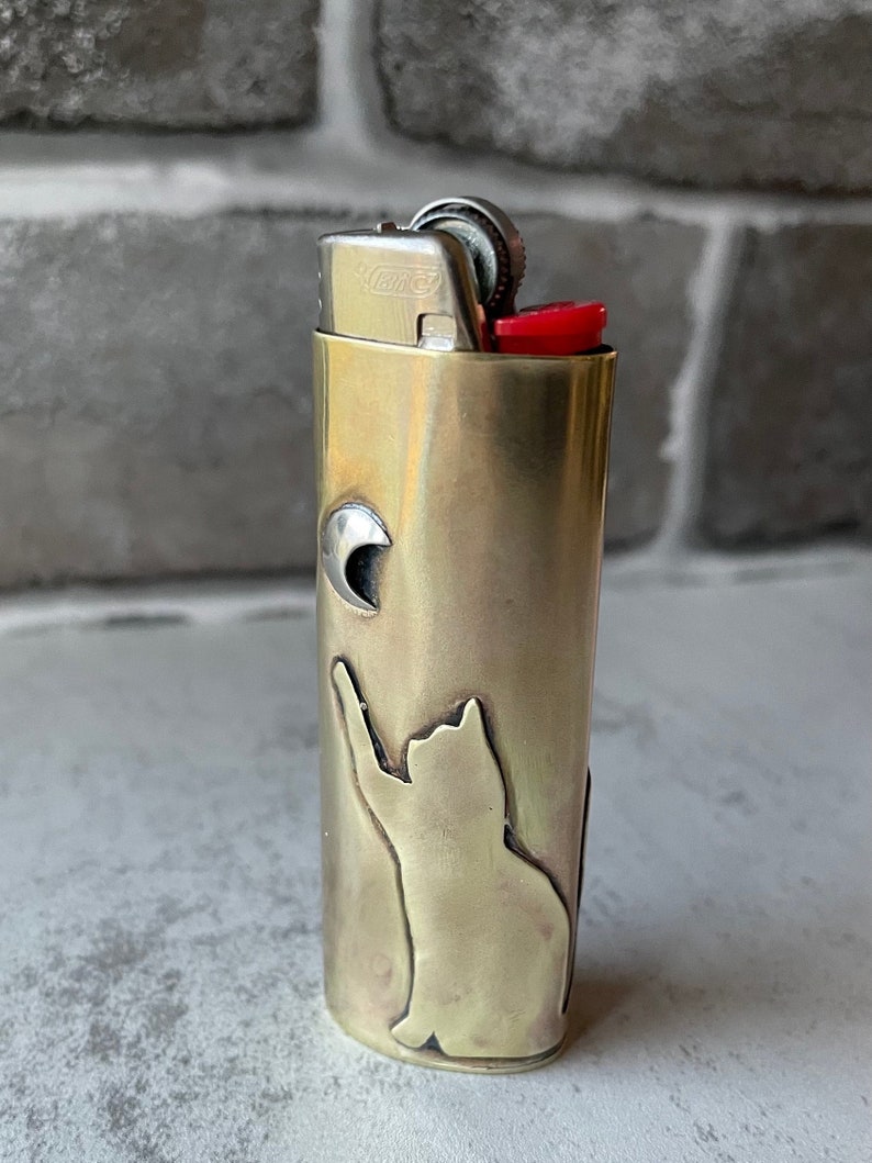 May include: A brass-colored lighter with a cat silhouette reaching for a crescent moon. The lighter has a red ignition button and a silver metal top. The design is etched into the brass casing.