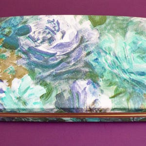May include: Rectangular box with a floral design in shades of blue, teal, and white. The box has a gold-colored metal trim and a rounded top. The background is a solid purple color.